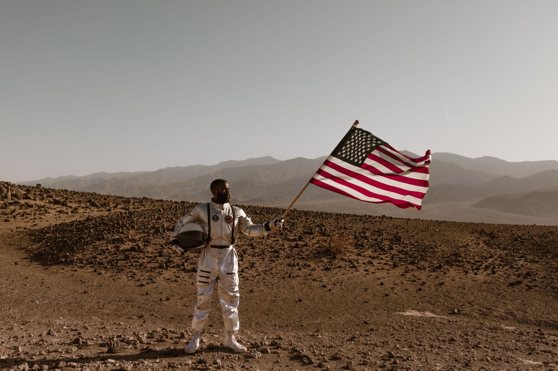An astronaut in a space suit stands in a desert landscape holding an American flag.