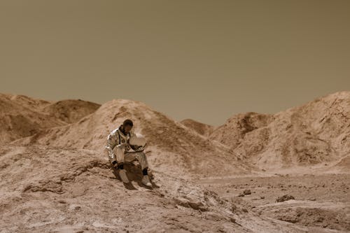 A Man in Space Suit Sitting on a Dry Rocky Ground