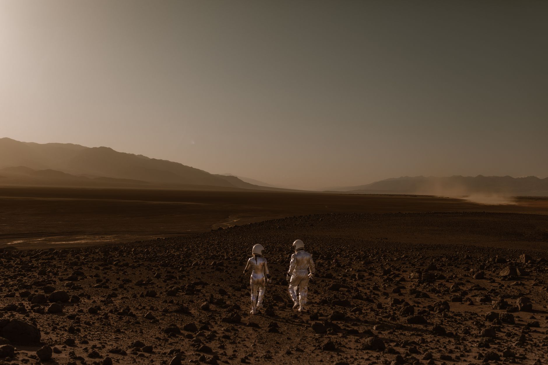Two astronauts in spacesuits walk across a Mars-like desert landscape during the day.