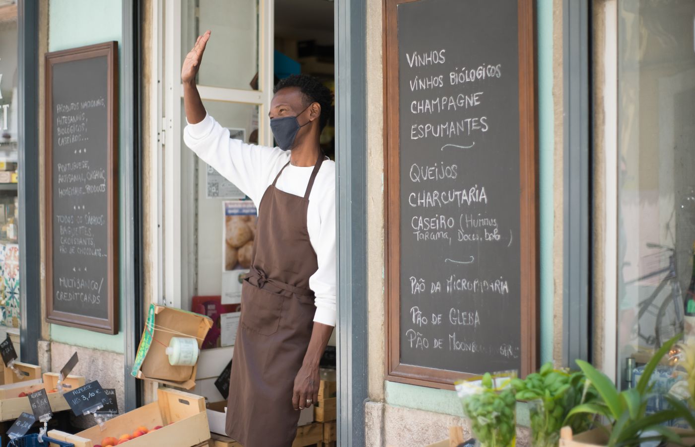 A grocery store clerk wearing an apron and face mask warmly waves from the storefront during pandemic times.