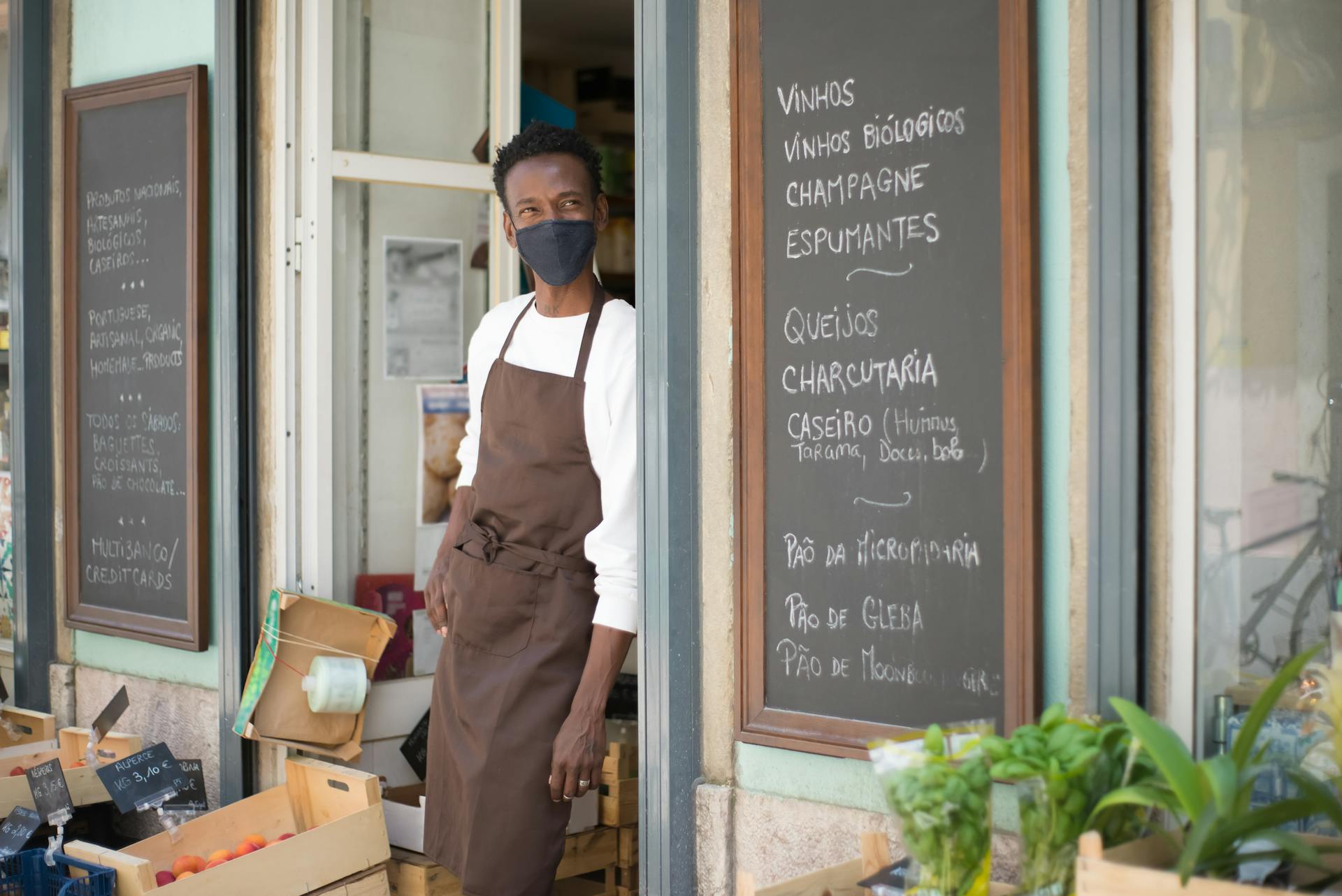 Portrait of a masked shopkeeper standing in a Portuguese store doorway with chalkboard menus.