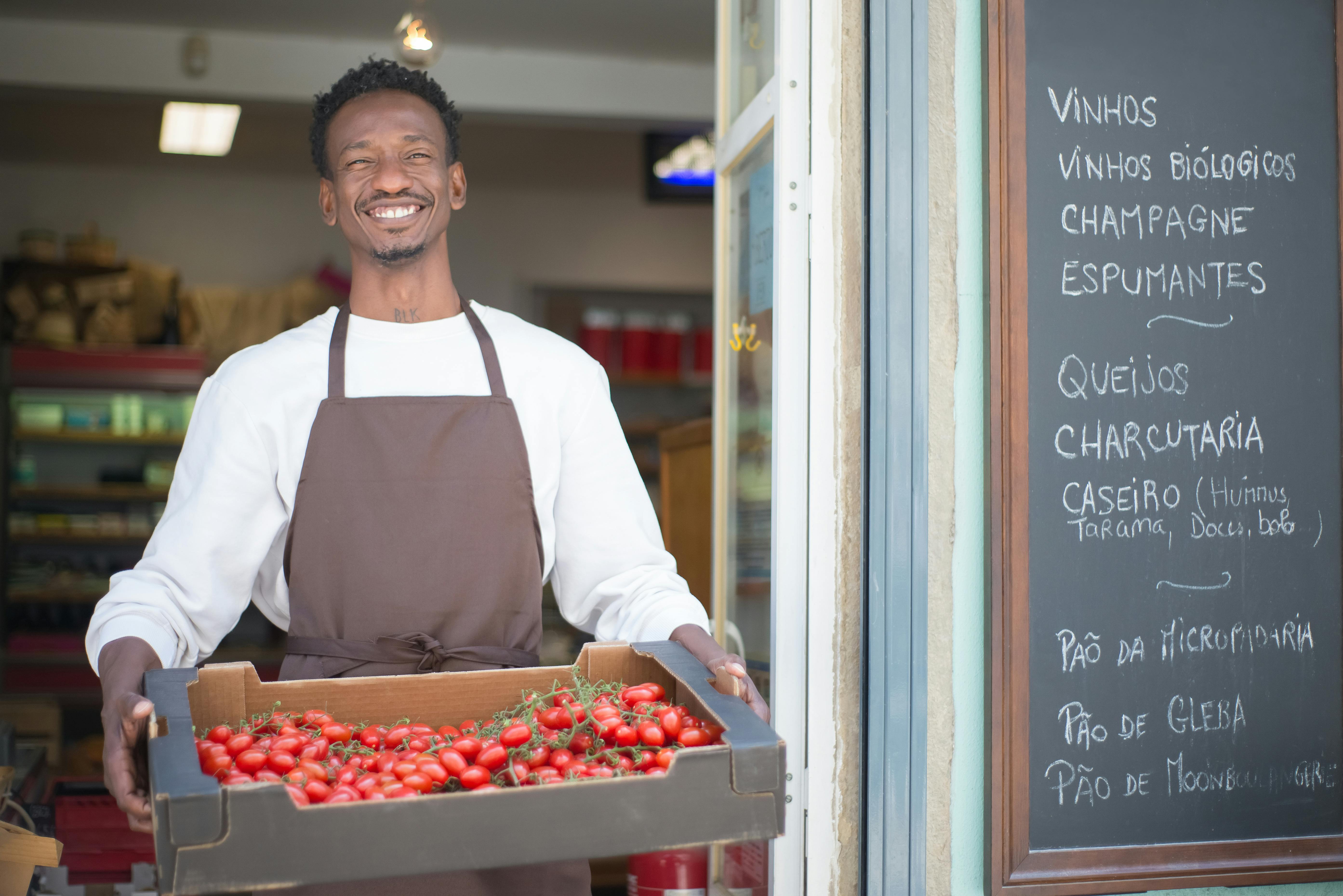 Smiling vendor standing outside a Portuguese store holding a box of ripe tomatoes. Perfect for grocery or food themes.