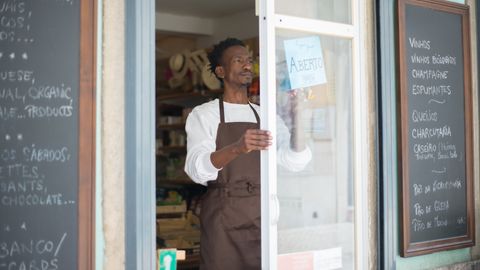 A man in an apron opens a grocery store door in Portugal, ready for business.