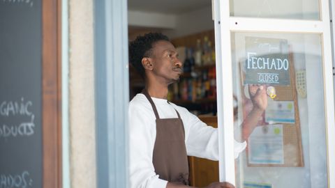 African man closing a store with a sign in Portuguese, wearing an apron.