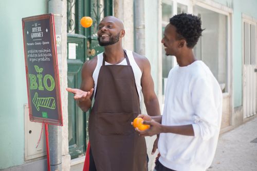Free Two men interacting by a grocery store, one juggling oranges with a sign in Portuguese nearby. Stock Photo
