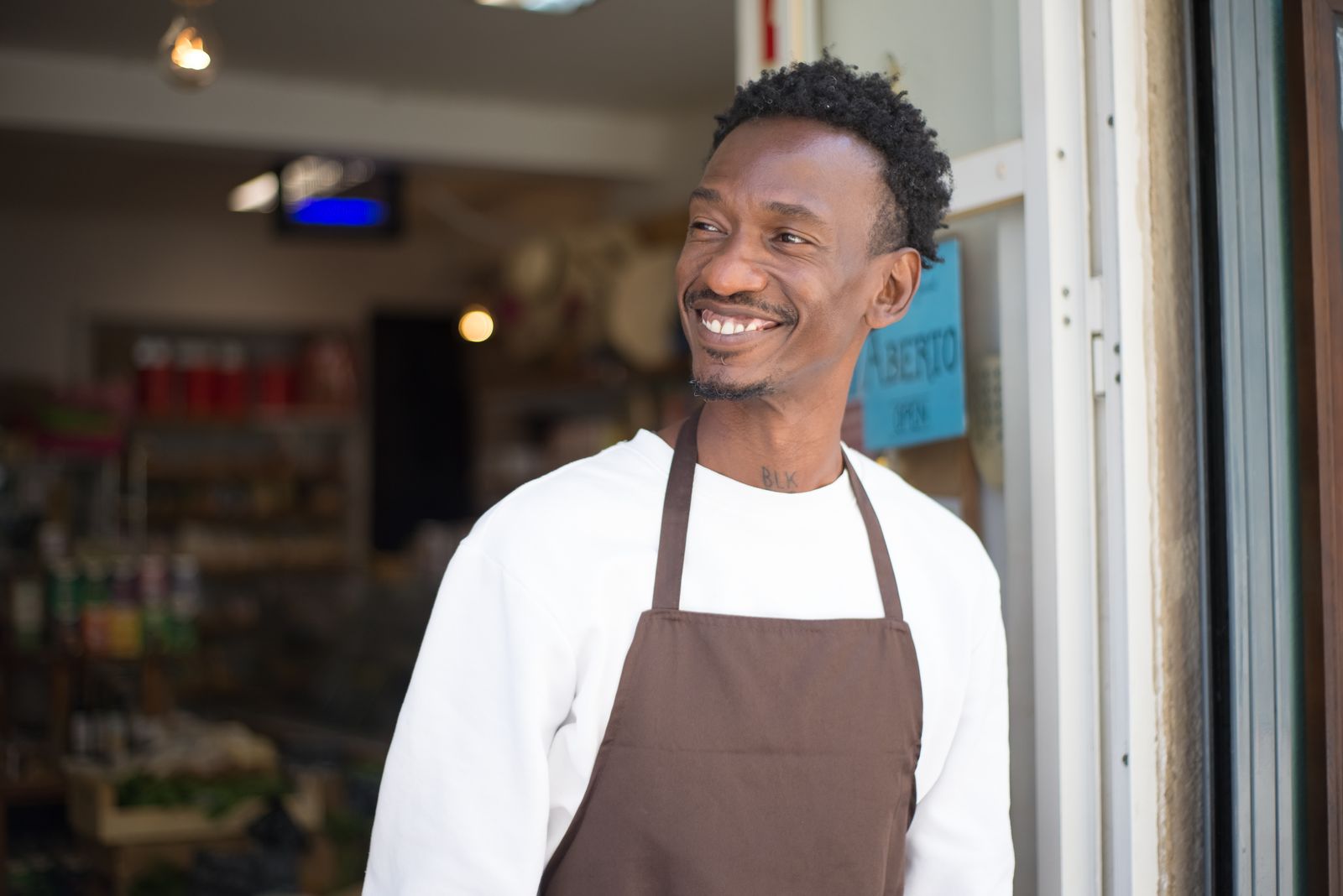 A smiling store owner in a brown apron standing proudly at the entrance of his shop.