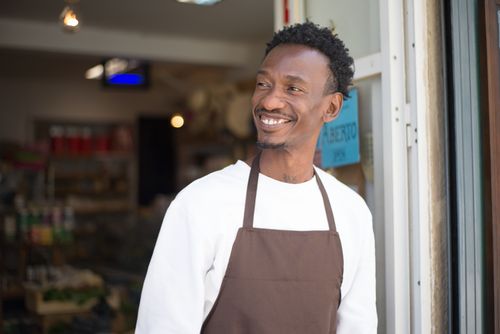 Free Smiling store owner in brown apron standing at entrance, welcoming customers. Stock Photo
