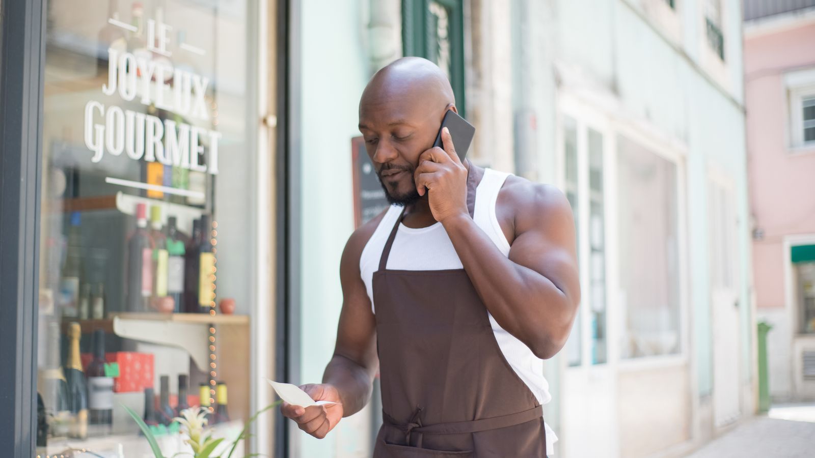 Man in apron making a business call outside a gourmet store in Portugal, checking notes.