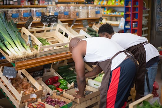 Two men in aprons arranging vegetables at a busy market stall indoors.