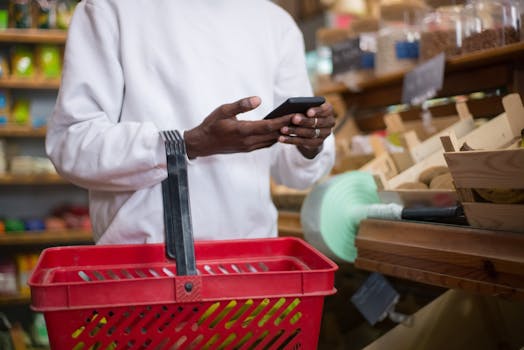 A man shops using a smartphone at a grocery store, holding a red basket filled with groceries.