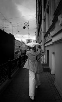 A woman walks under an umbrella on a rainy city sidewalk in black and white.
