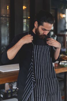 A bearded chef adjusting his navy striped apron in a restaurant kitchen setting.