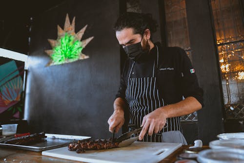 A Man in Black Long Sleeves Wearing Face Mask while Slicing a Steak