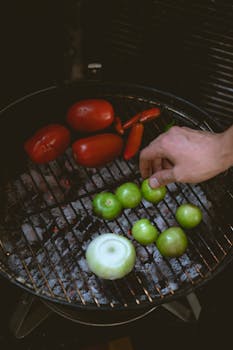 Hand arranging tomatoes, chilies, and onions on a charcoal grill for salsa.