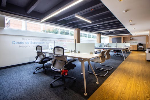 A well-lit, modern office space featuring ergonomic chairs, tables, and glass dividers.