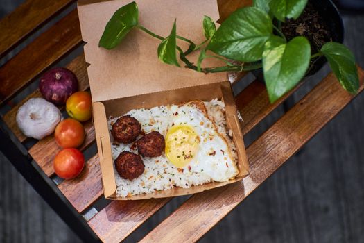 A delicious breakfast box of rice, fried egg, and falafel on a rustic wooden table.