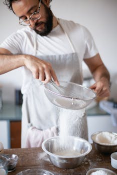 A focused male baker sifting flour into a metal bowl in a cozy kitchen setting.
