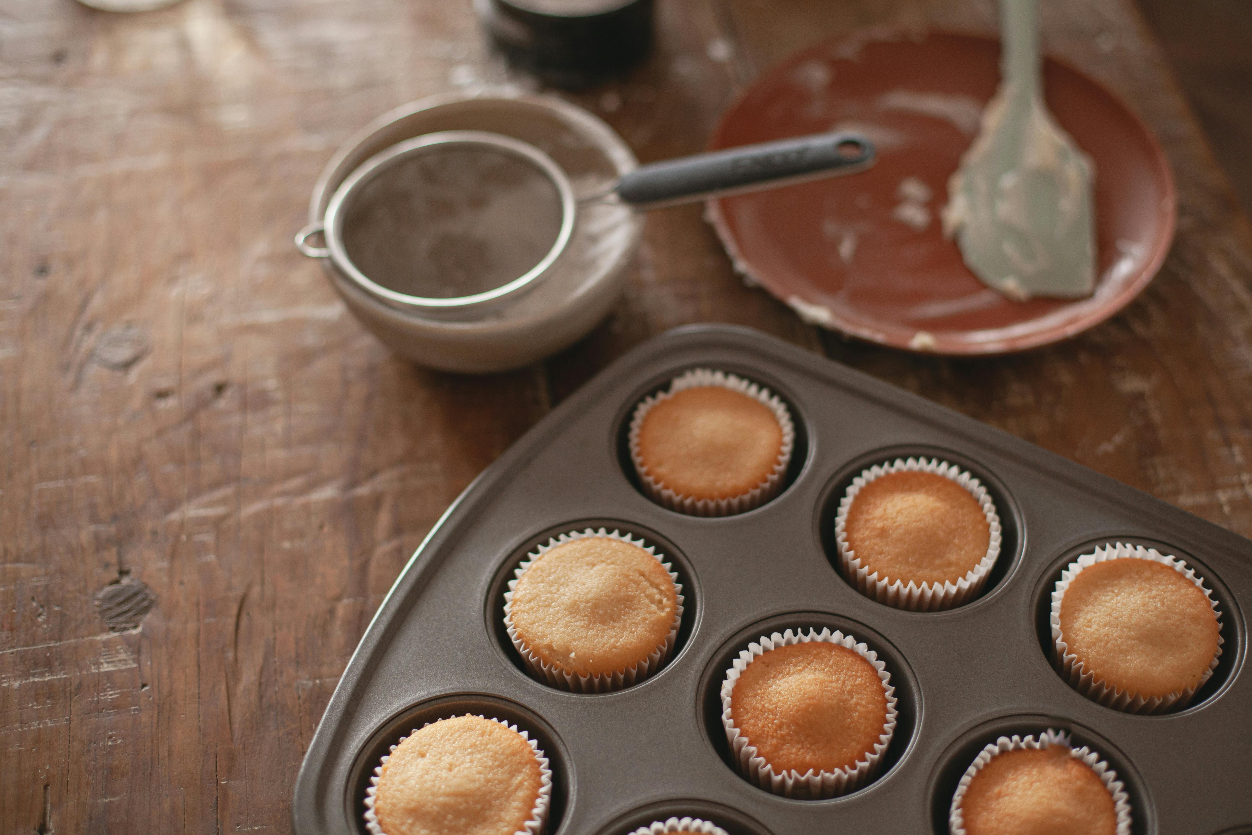 Warm cupcakes sit fresh from the oven in a muffin tin on a rustic table setting.