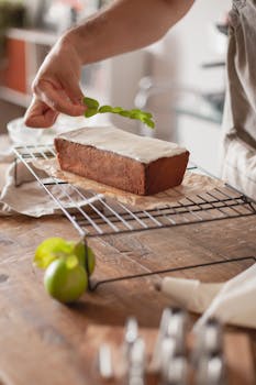 Close-up of a hand placing mint leaves on glazed cake in kitchen setting.