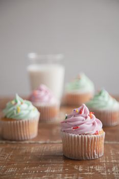 Close-up of colorful frosted cupcakes on a wooden table with a glass of milk in the background.