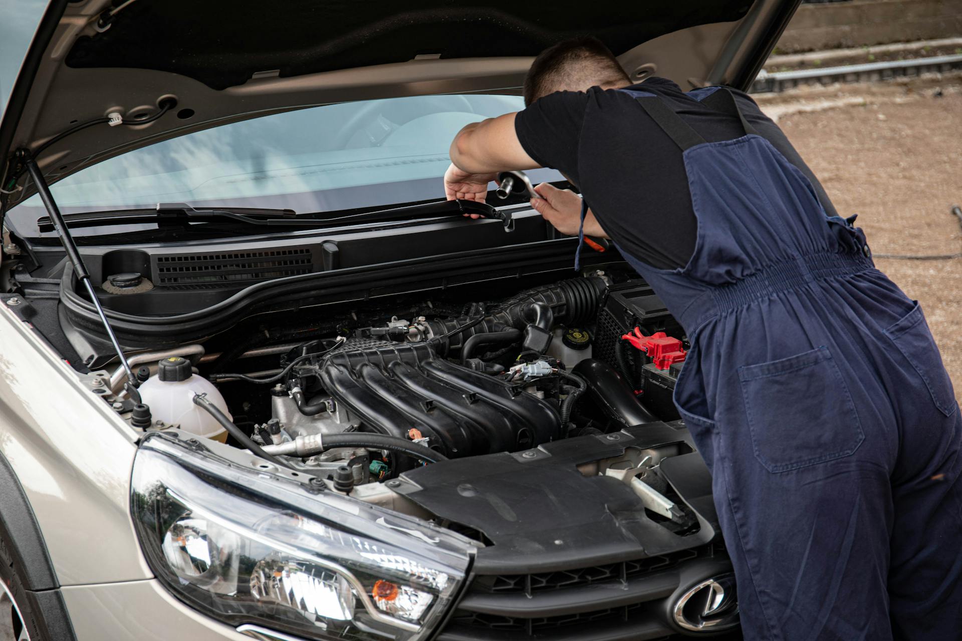 A mechanic in blue overalls works on a car engine in an outdoor setting.