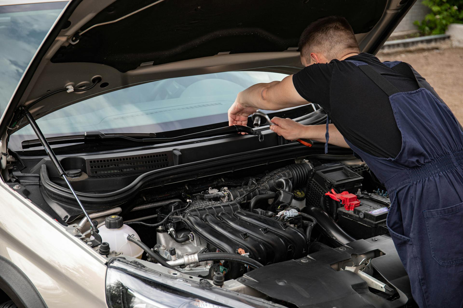 A mechanic uses tools to repair a car engine outdoors in daylight.