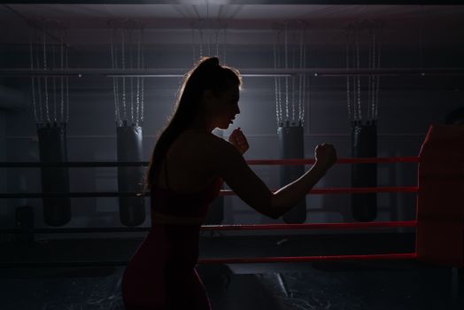 Silhouette of a woman boxer practicing in the gym with punching bags in the background.