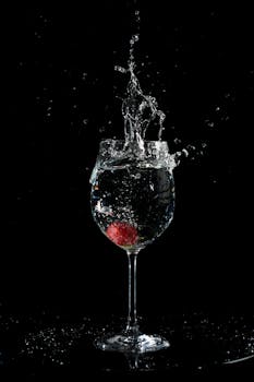 A red fruit causing a splash in a wine glass against a black background.