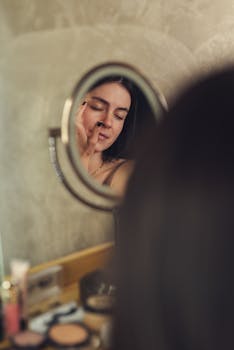 A woman with eyes closed, applying makeup in front of a mirror for a serene, morning routine moment.