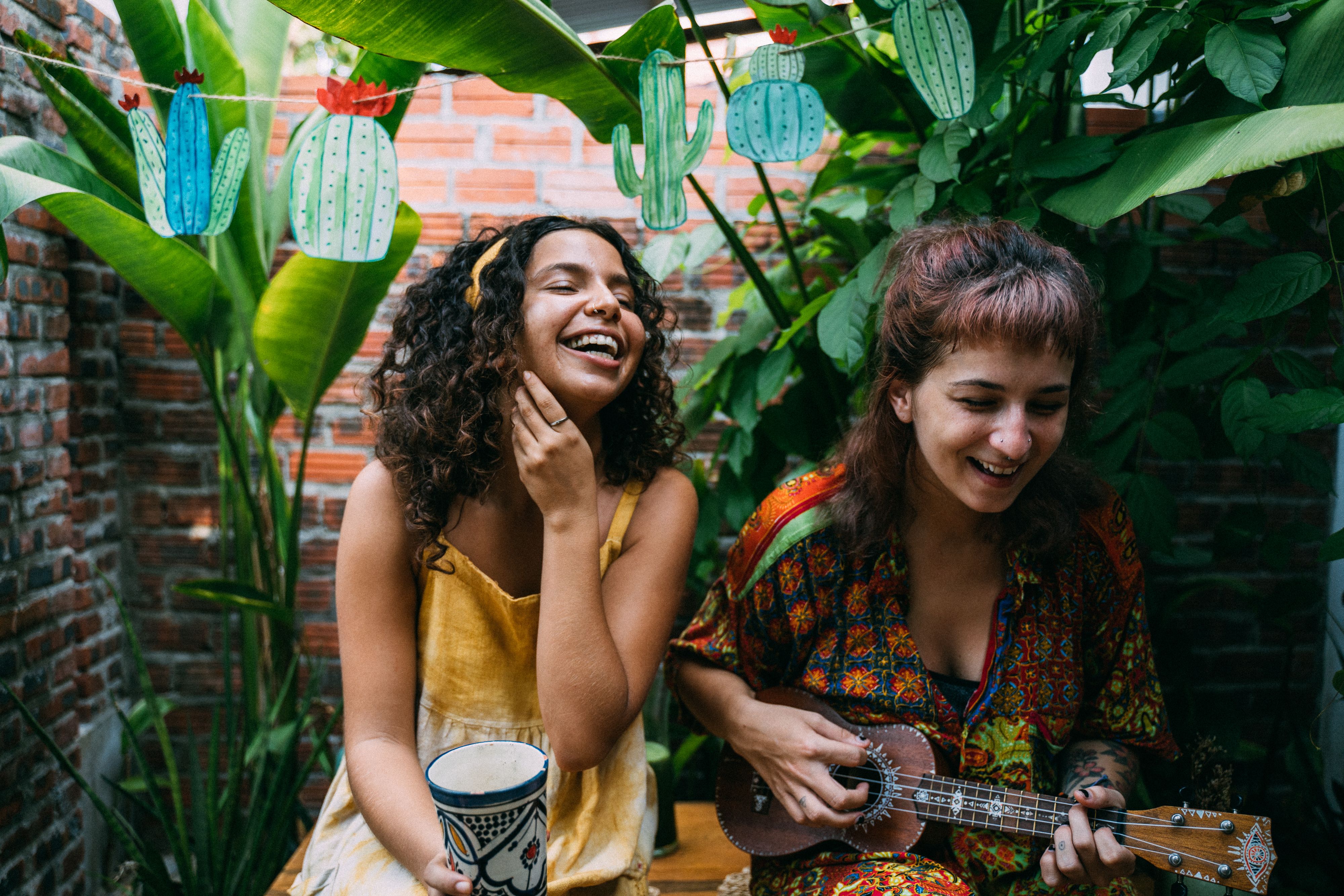Two women laughing and playing music in an outdoor garden setting with lush greenery.