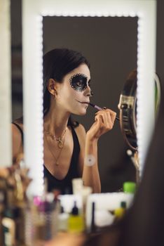 Woman in mirror applying Catrina makeup for Day of the Dead celebration indoors.