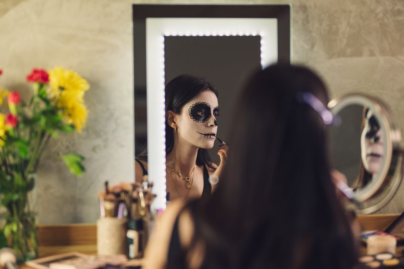 Woman applying Catrina makeup for Day of the Dead in front of a mirror adorned with flowers.