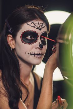 A woman carefully applies traditional Catrina makeup for Dia de los Muertos, featuring intricate designs.