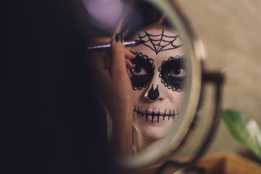 Woman in front of mirror applying Day of the Dead face paint for Halloween celebration.