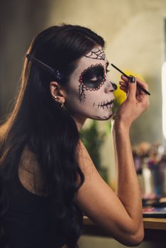 A Hispanic woman applying Day of the Dead makeup indoors during Halloween.