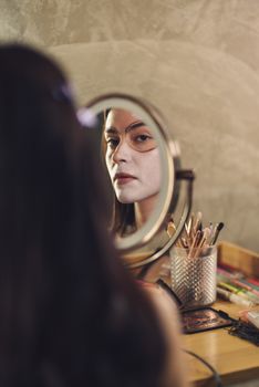 A woman applying Catrina makeup for Halloween while looking in the mirror indoors.