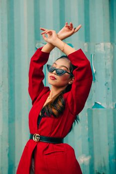 Stylish woman in red coat posing with raised arms and sunglasses against a blue backdrop.