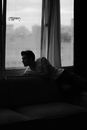 Black and white of pensive young male in casual clothes sitting on comfortable sofa near window while looking away in apartment