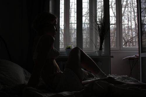 Free Silhouette of a woman sitting by a window in a dimly lit bedroom, creating a moody atmosphere. Stock Photo