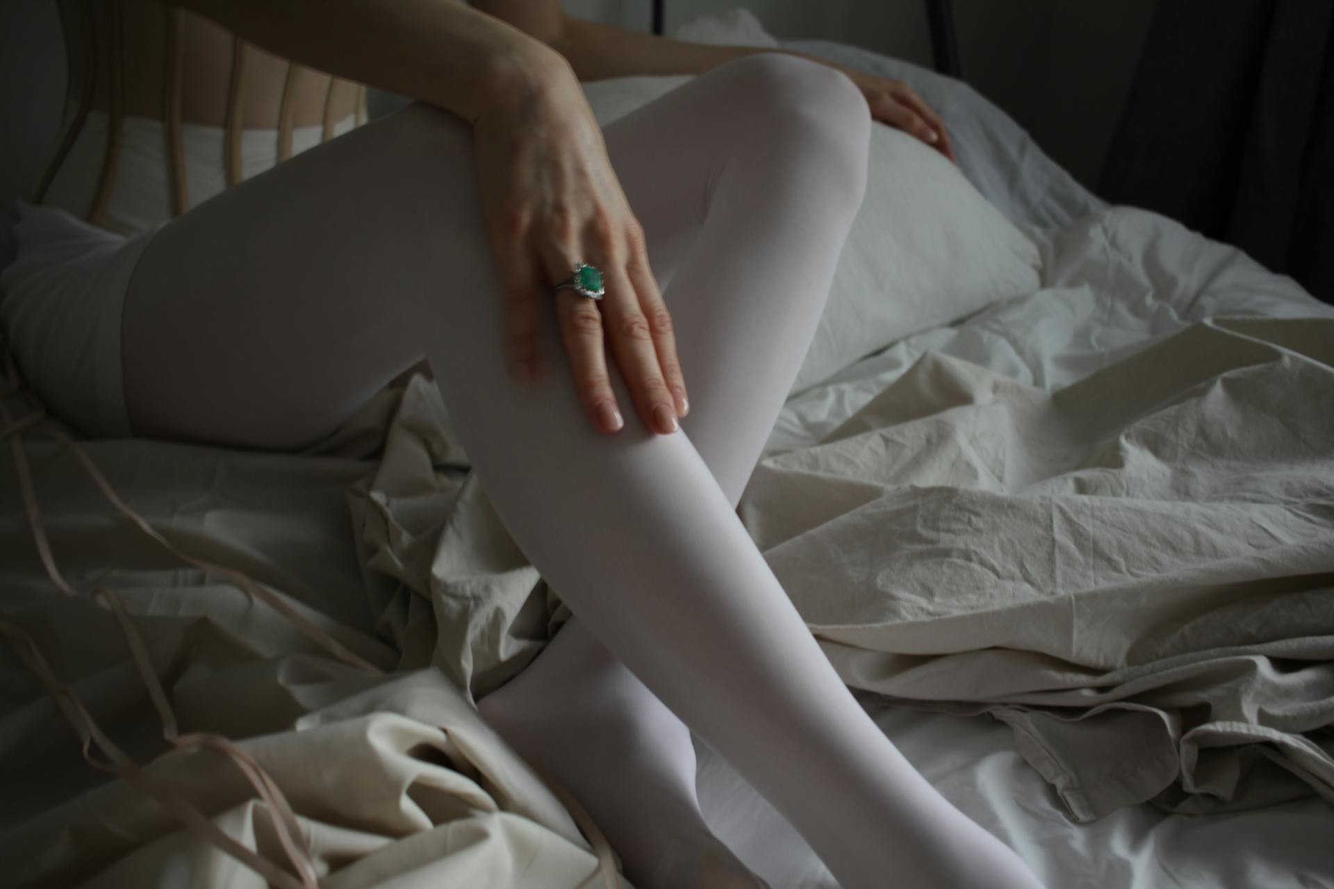 Close-up of a woman's legs in white stockings with an emerald ring on a white bed.