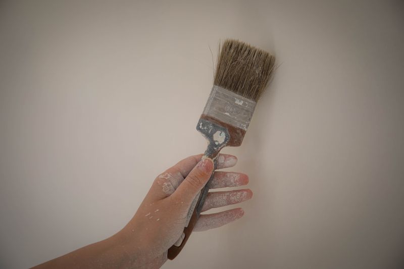 A hand holds a paintbrush with white paint against a plain wall, showcasing DIY home improvement.