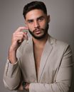 Masculine man in gray jacket with beard and modern haircut touching cheek while looking at camera