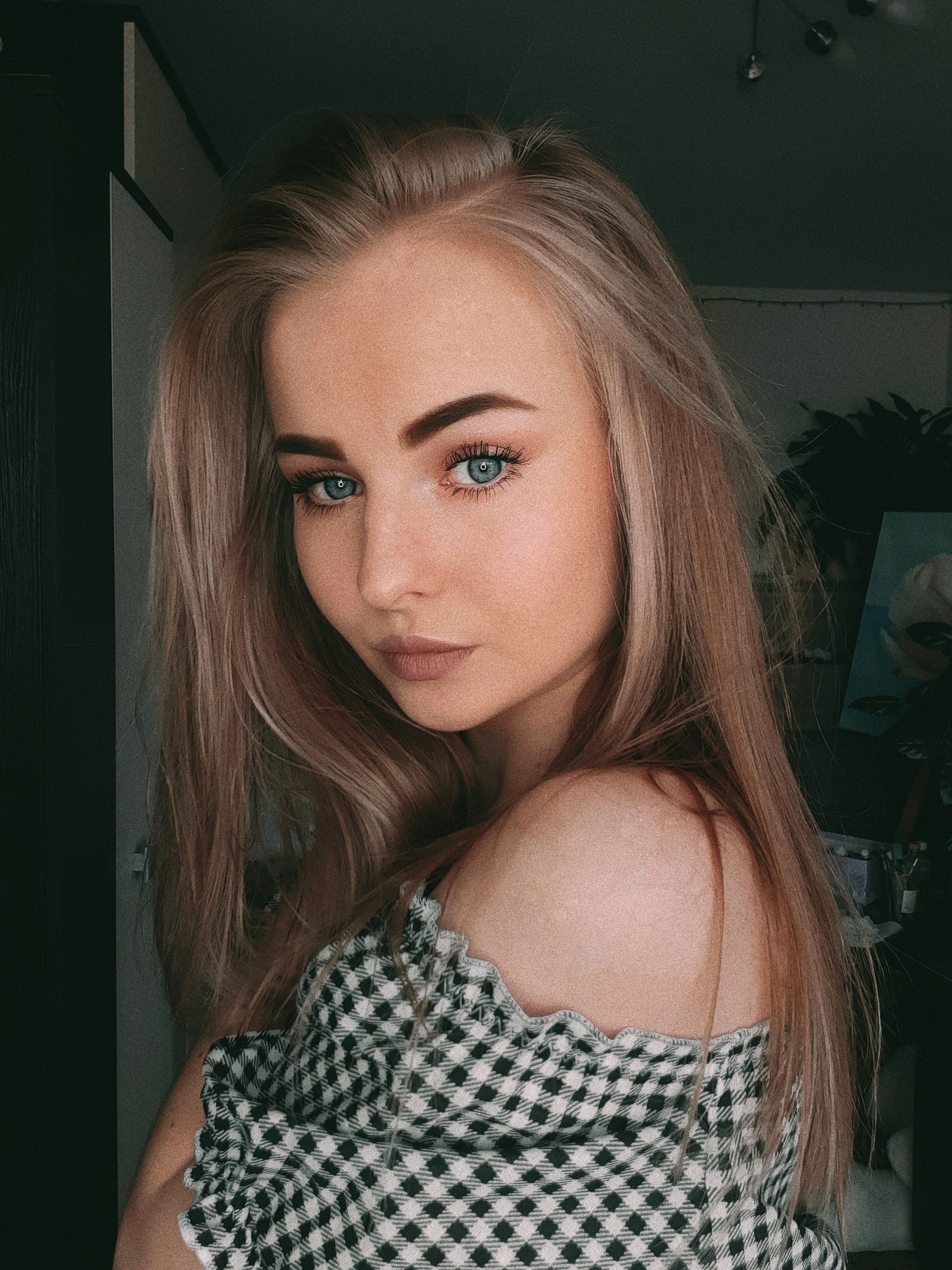 Close-up portrait of a young woman with blonde hair posing indoors in a checkered top.