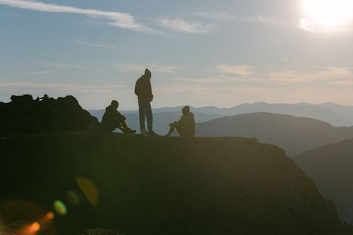 Free Three friends silhouetted on a mountain ridge under a glowing sun. Capturing tranquility and adventure. Stock Photo
