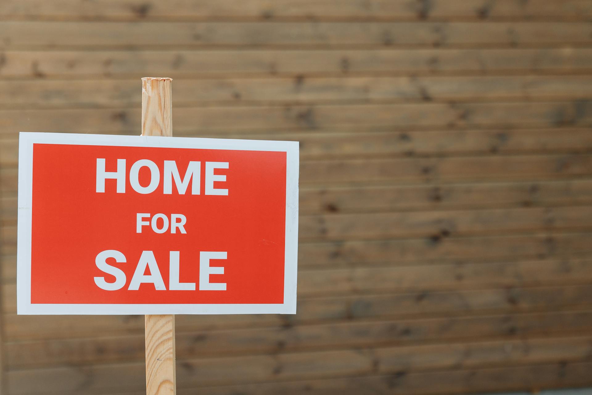 Close-up of a red home for sale sign against a wooden backdrop, ideal for real estate use.