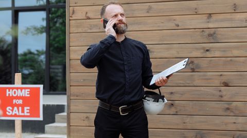 Real estate agent making a phone call while holding documents near home for sale sign.