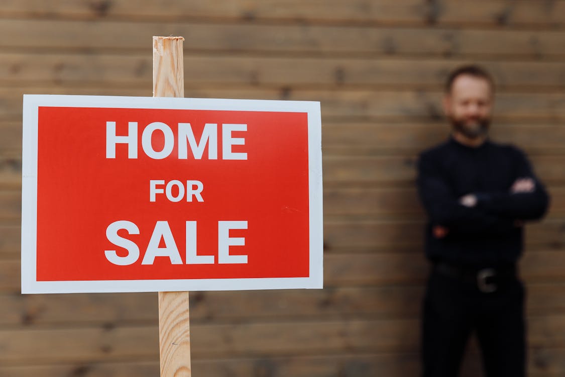 Free A confident real estate agent stands by a home for sale sign against a wooden wall. Stock Photo