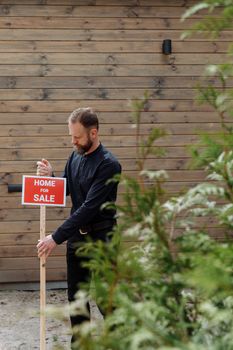 A real estate agent placing a 'home for sale' sign outside a house in a natural setting.