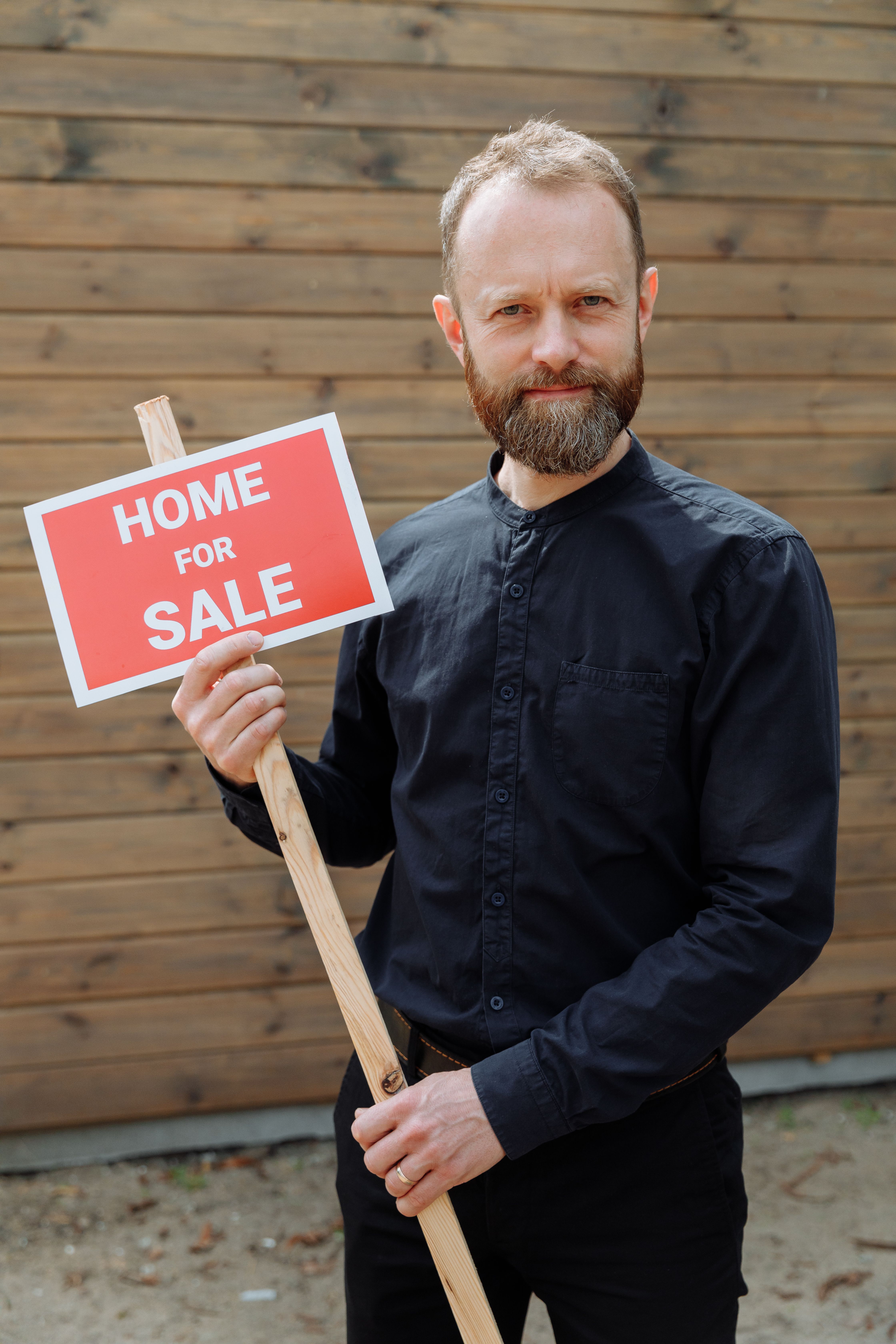 Confident real estate agent with a beard holding a 'Home for Sale' sign outdoors.