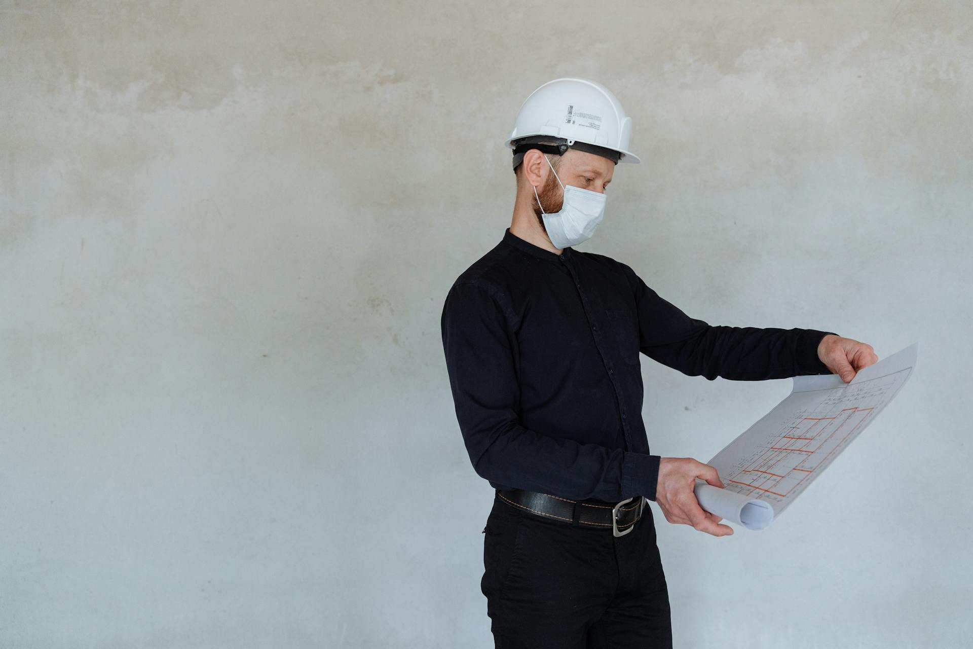 Architect in safety helmet and mask reviewing building plans indoors.